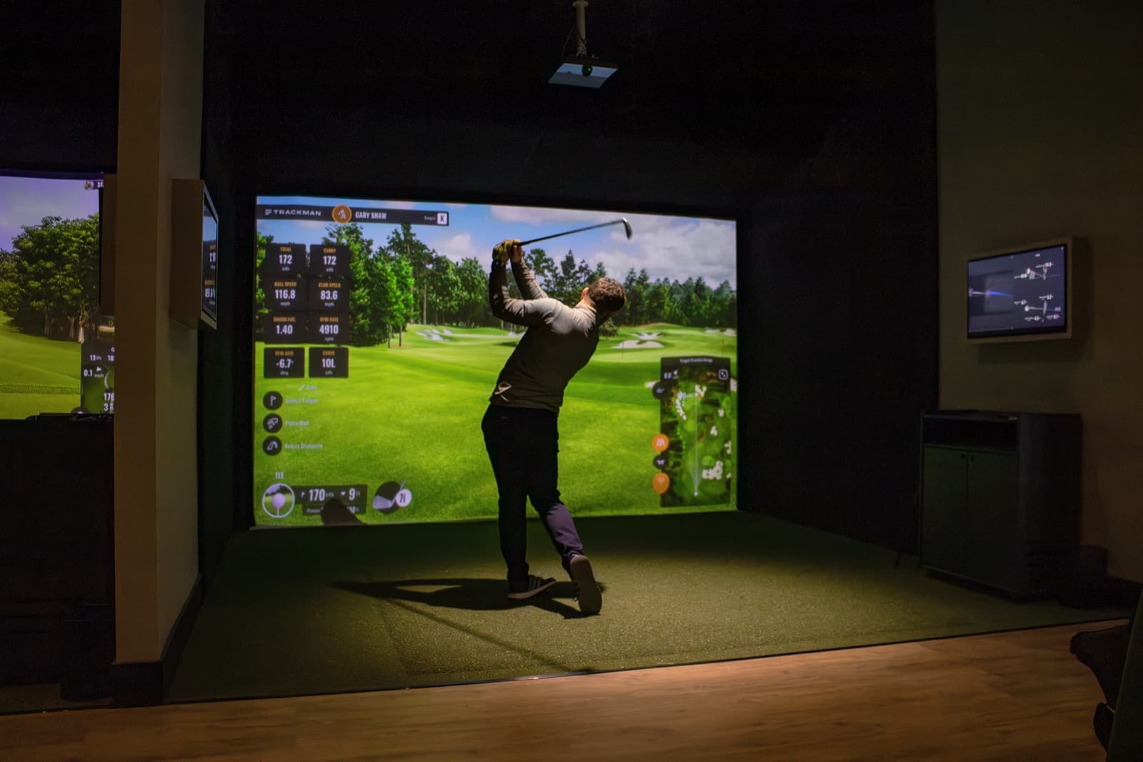 A golfer mid-swing in a Caddi Club bay, a TrackMan course on the big screen