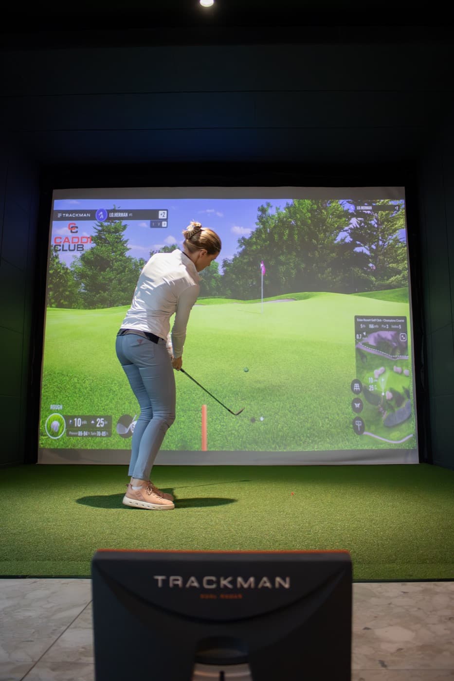 A golfer mid-swing in a Caddi Club bay with the TrackMan unit in the foreground